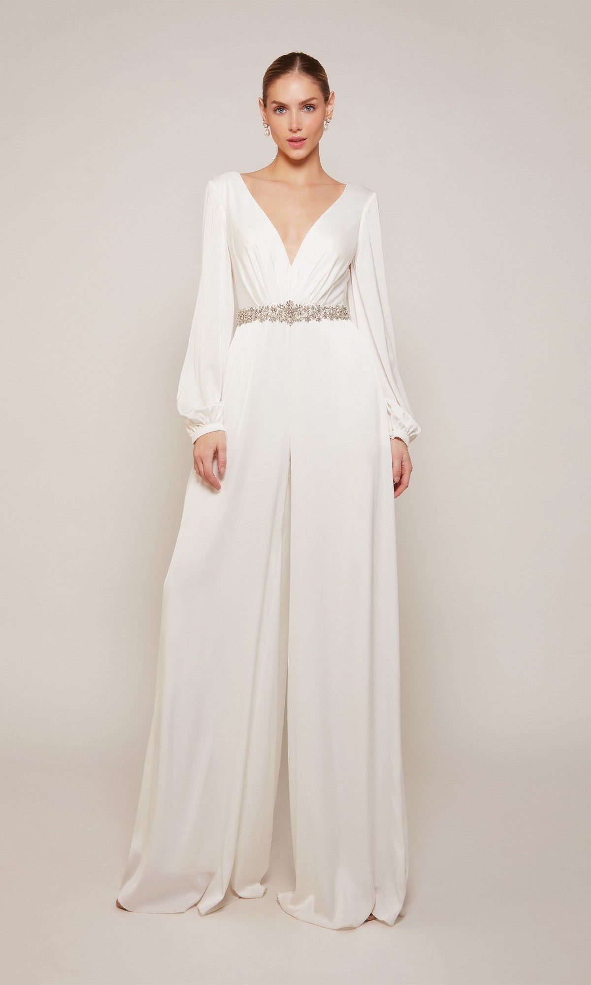 A woman models the Alyce 70031, a white satin-chiffon formal jumpsuit with long sleeves, wide legs, a deep V-neckline, and jeweled belt, set against a plain light background.